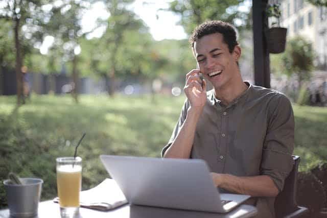 A man talking on the phone while sitting at an outdoor table with a laptop.